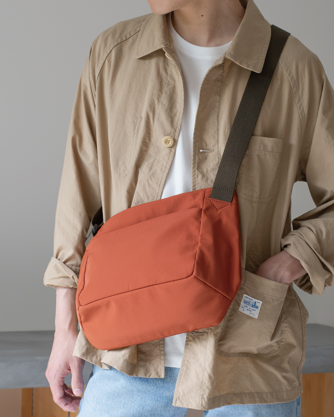 男性がオレンジ色のメンズショルダーバッグを肩にかけている全身ショット。カジュアルな服装に合うデザイン。  
