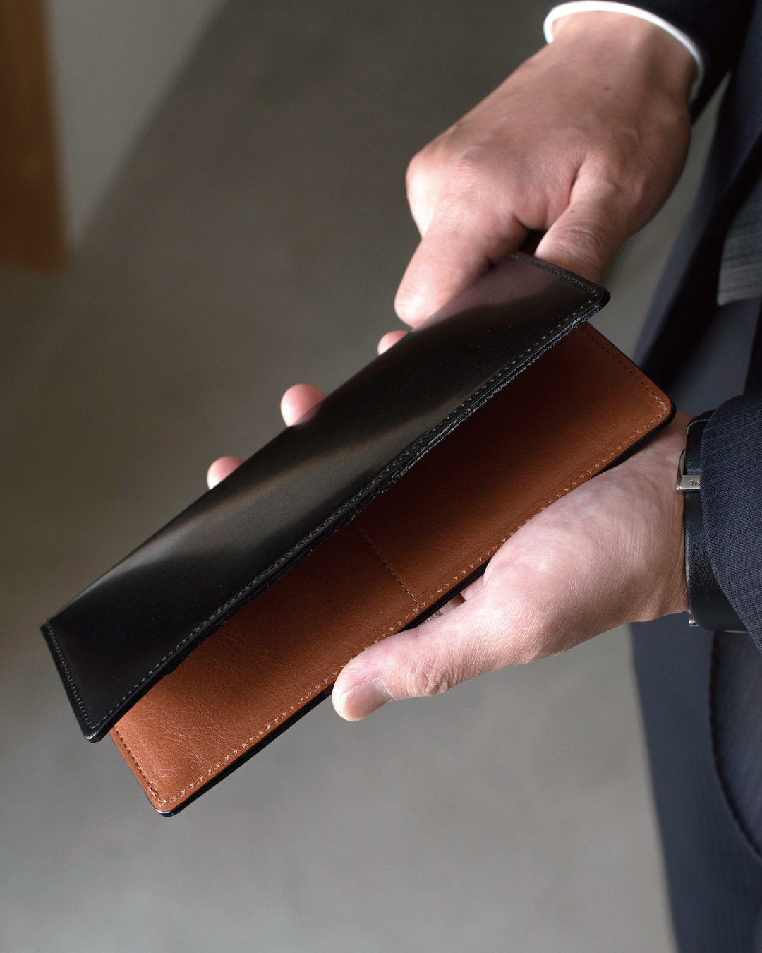 スーツを着た男性がブラックのコードバン長財布を手に持っている様子。ビジネスシーンに馴染む落ち着いた高級感と、財布を開いた際の内装のブラウンのコントラストを示している。