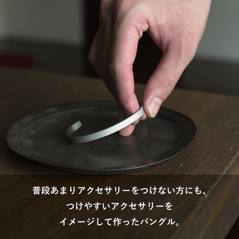 普段アクセサリーをつけない方にも取り入れやすい、シンプルなTVRT 真鍮バングルを指でつまんで黒い器に置く様子