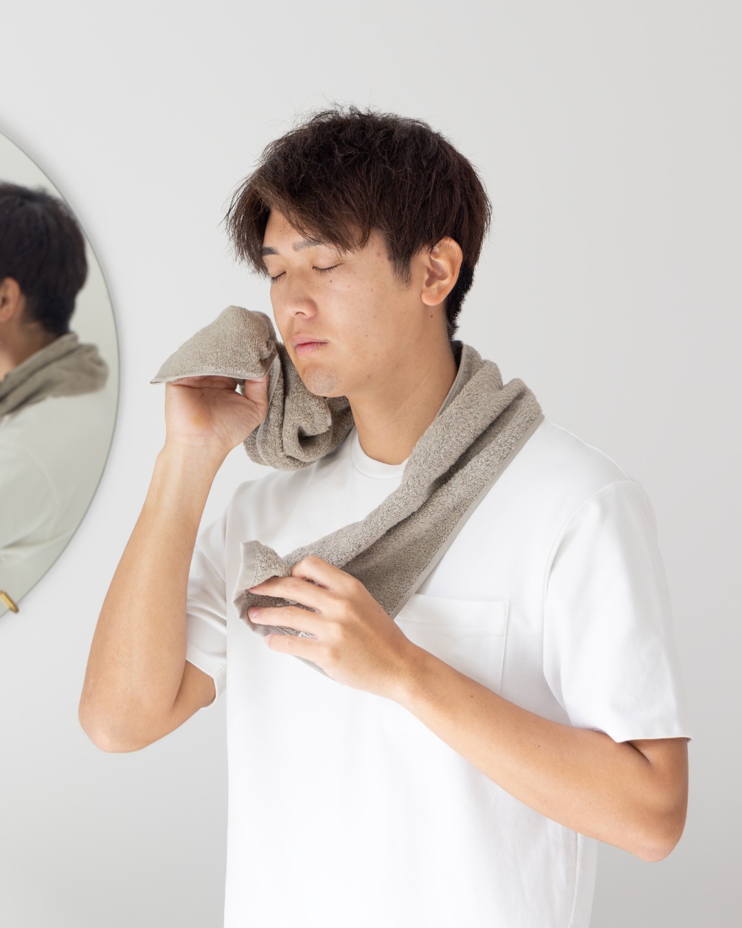 トープカラーのちょうどいいマフラータオルを首にかけて洗顔後の顔を拭く男性。鏡のあるバスルームシーンで柔らかな肌触りと日常使いの快適さを表現。