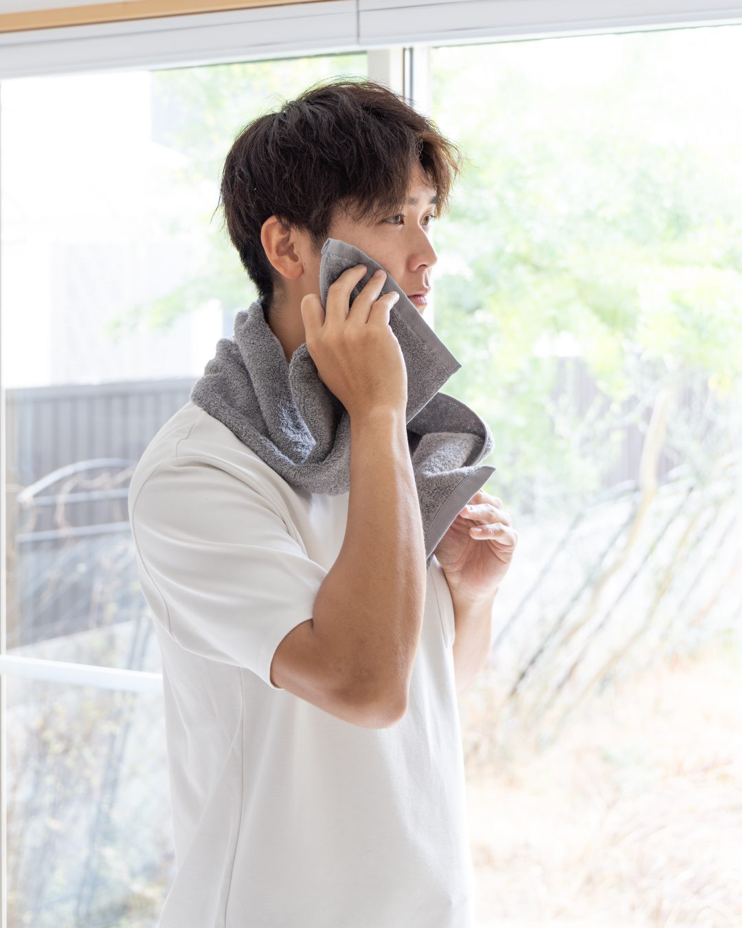 グレーのちょうどいいマフラータオルで顔を拭く男性の横顔。窓際の自然光が降り注ぐ爽やかな朝のシーンで高吸水性の今治タオルの使い心地を演出。