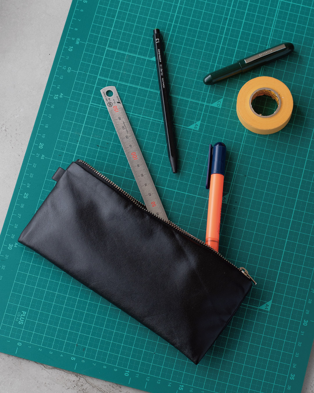カッティングマット上でFOLMAレザーポーチPENから定規・ペン・マーカーが飛び出したフラットレイ。スリムな横長シルエットと艶やかなブラックナッパーレザーの質感を俯瞰で表現。