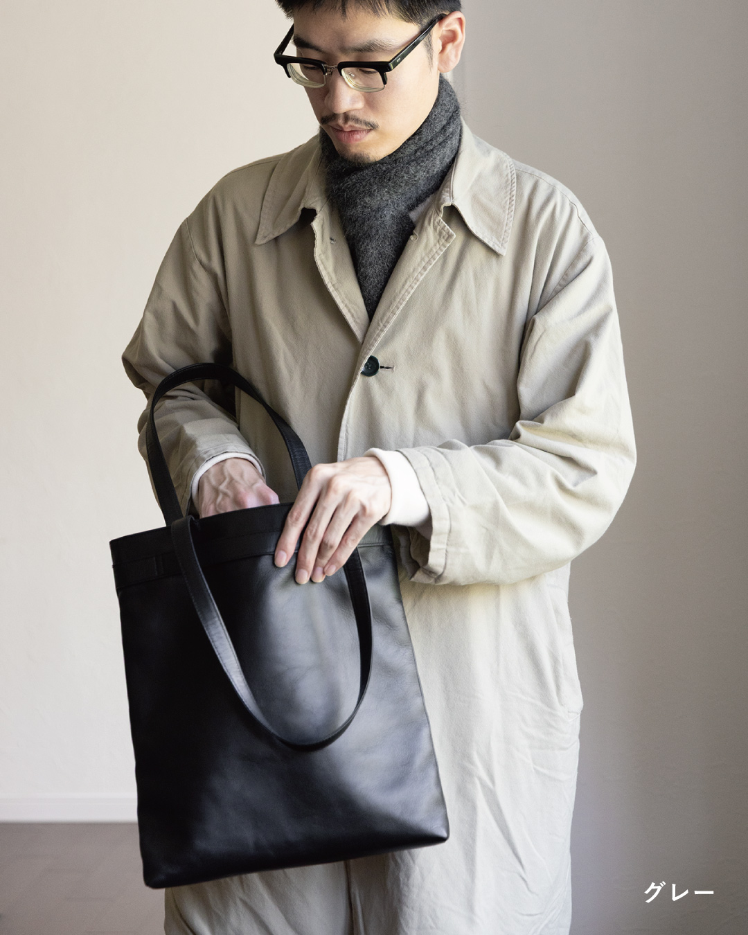 レザートートの中を確かめる男性が身につけた、グレーの3秒マフラー キッドモヘヤとベージュコートの軽やかな重ね着をとらえた日常のワンシーン。
