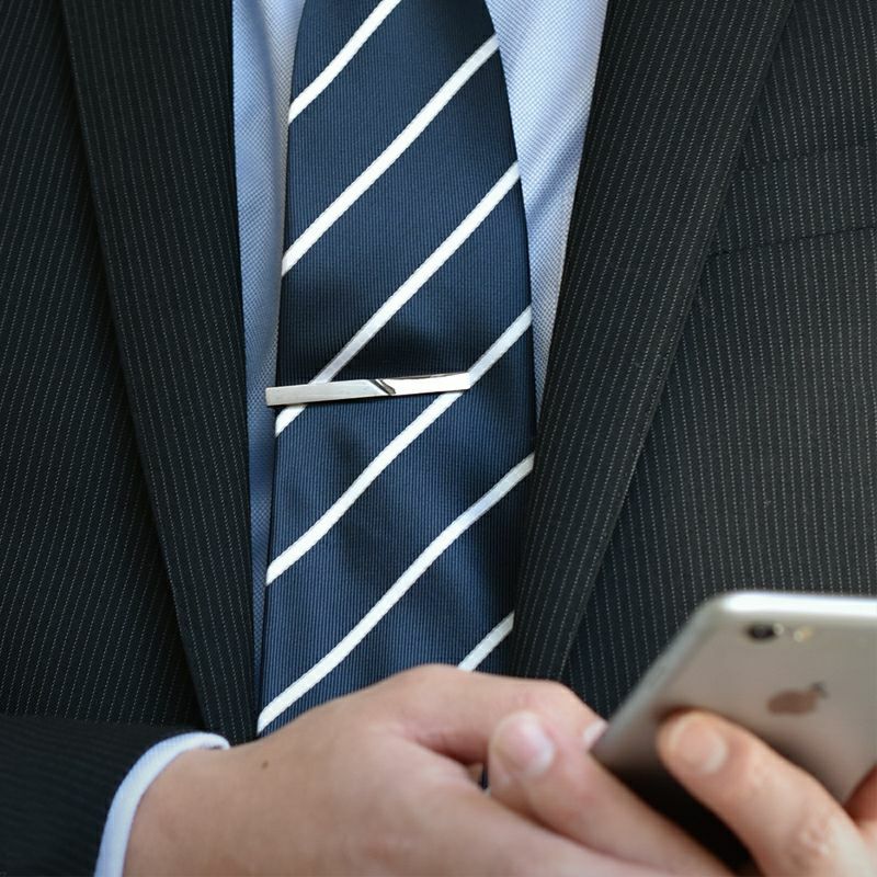 ストライプ柄のネイビーのネクタイに着用されたシルバーのジオメトリックネクタイピンとスマートフォンを持つ男性。